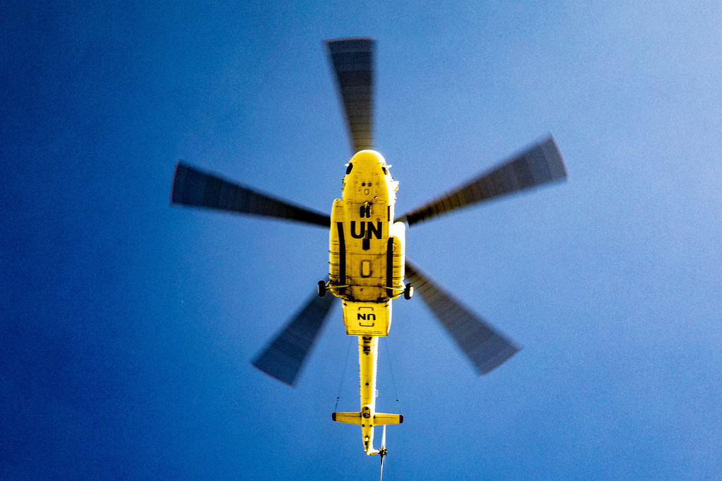 Um helicóptero amarelo das Nações Unidas com as marcas "ONU" voa sobre a aldeia de Ogossagou, no centro do Mali, contra um céu azul claro.