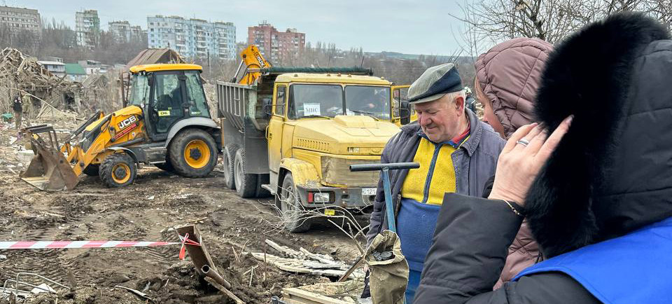 Украинские унтер-офицеры работают на месте недавнего обстрела в Днепре, на заднем плане — тяжелая техника и обломки.