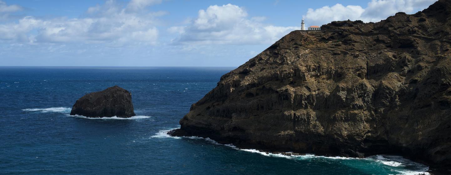 Um litoral rochoso em Cabo Verde, com um farol no alto de uma falésia com vista para o oceano e uma formação rochosa na água.