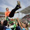 Un portero masculino con una camiseta naranja es levantado en el aire por sus compañeros de equipo en celebración en un campo de fútbol.