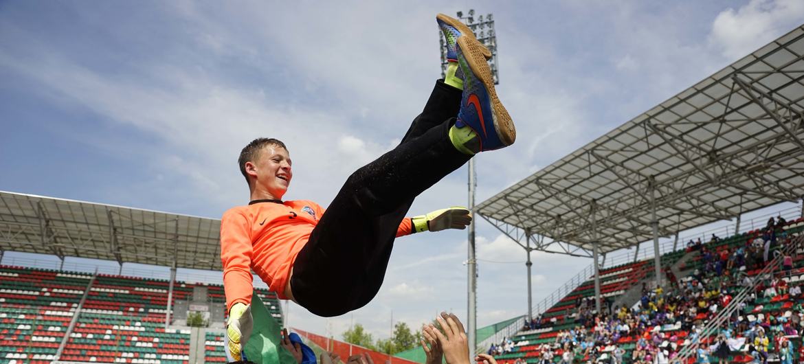 Un portero masculino con una camiseta naranja es levantado en el aire por sus compañeros de equipo en celebración en un campo de fútbol.
