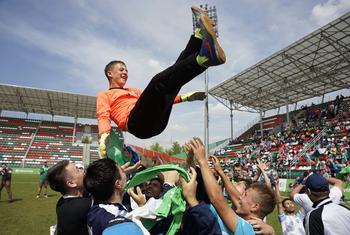 Un portero masculino con una camiseta naranja es levantado en el aire por sus compañeros de equipo en celebración en un campo de fútbol.