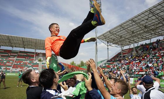 Un portero masculino con una camiseta naranja es levantado en el aire por sus compañeros de equipo en celebración en un campo de fútbol.