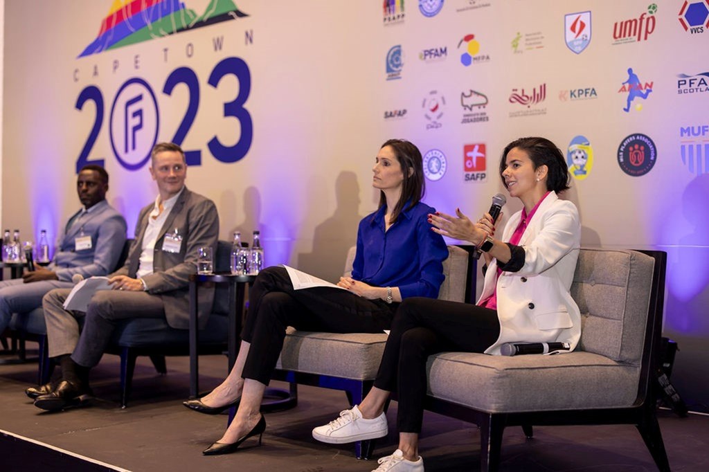 La exfutbolista Camila García habla ante un micrófono en el escenario de la Asamblea General de FIFPRO Ciudad del Cabo 2023, sentada junto a otros panelistas.
