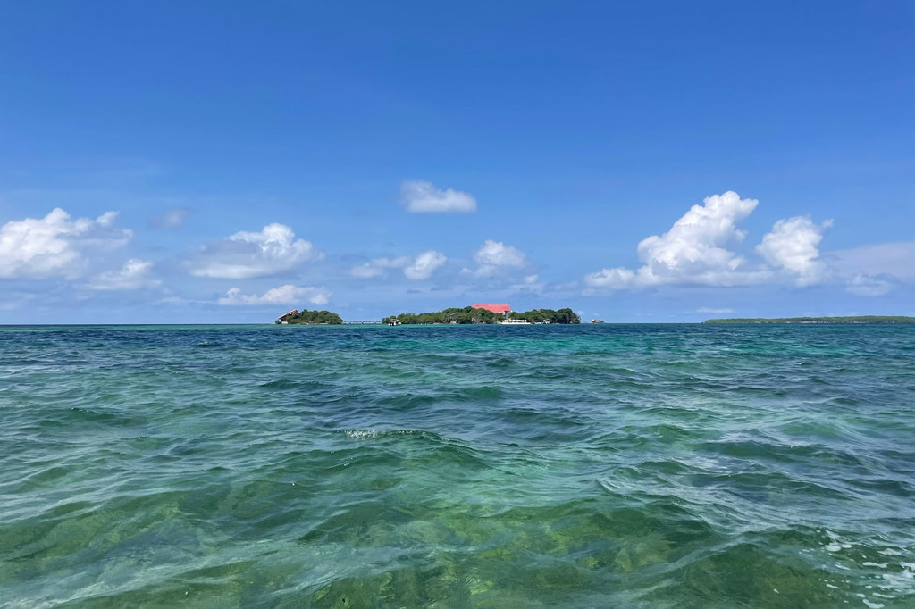 Una vista de las aguas turquesas del Mar Caribe con varias islas pequeñas en la distancia bajo un cielo azul con nubes blancas.