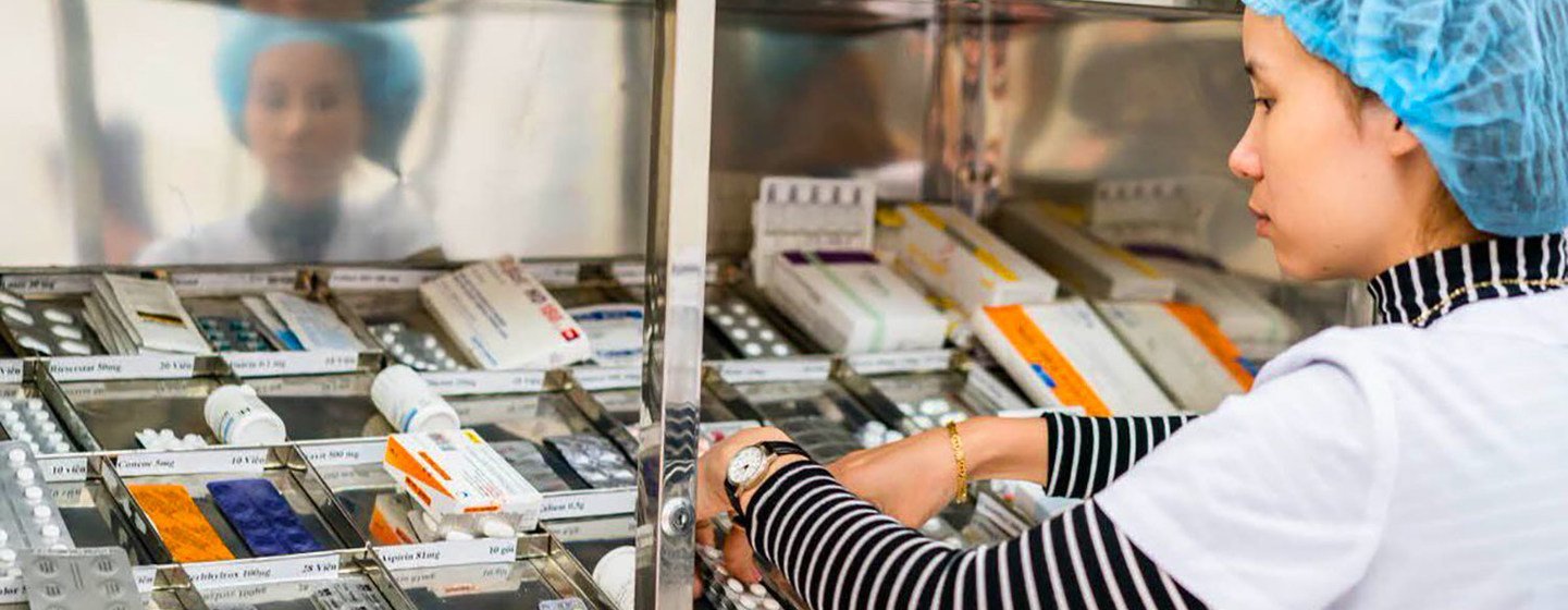 Dans la pharmacie d'un hôpital, une pharmacienne portant une charlotte range des médicaments dans une armoire métallique.
