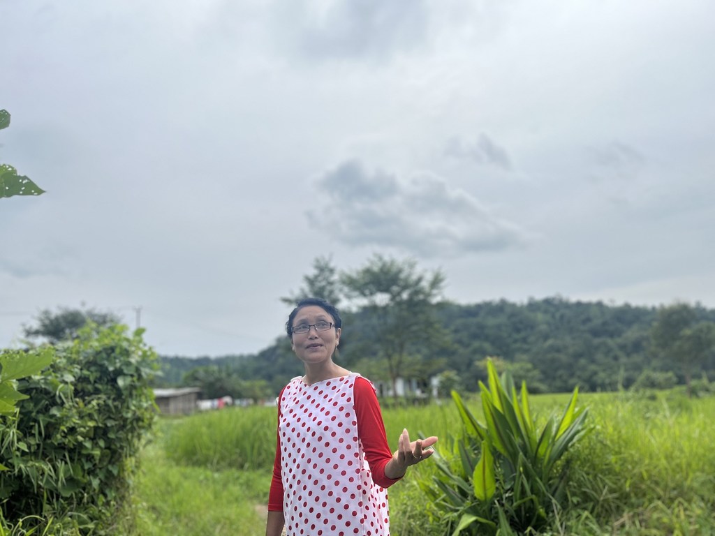 आयशा भारत के मेघालय में एक हरे-भरे मैदान में खड़ी हैं, उन्होंने लाल और सफेद रंग की पोल्का डॉट वाली ड्रेस पहनी हुई है और अपने हाथ से इशारा कर रही हैं।
