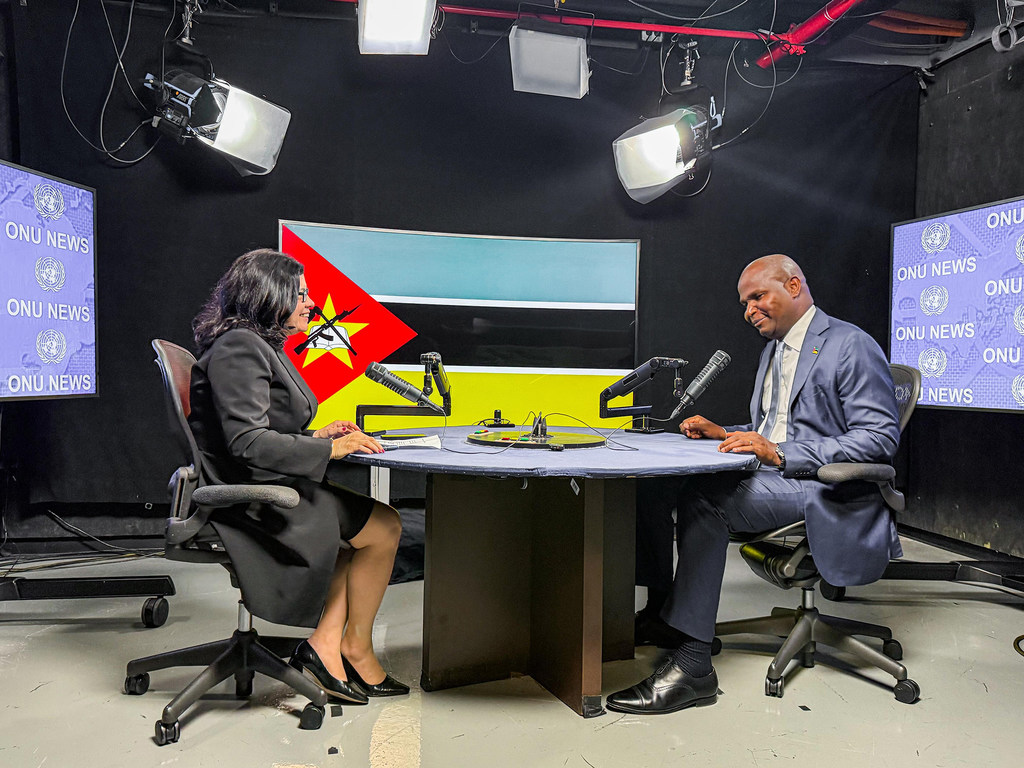 Uma repórter entrevista um homem em um estúdio com a marca ONU News e as bandeiras das Nações Unidas e de Moçambique ao fundo.