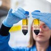 A scientist in a lab coat and gloves holds two test tubes containing yellow and red liquid samples.