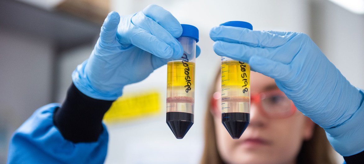 A scientist in a lab coat and gloves holds two test tubes containing yellow and red liquid samples.