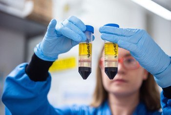 A scientist in a lab coat and gloves holds two test tubes containing yellow and red liquid samples.
