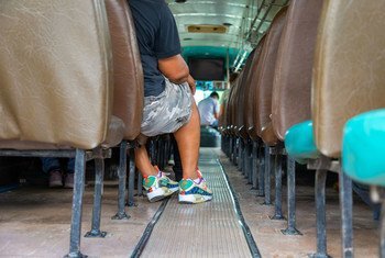Interior view of a bus with a passenger sitting on a brown seat, wearing camouflage shorts and colorful sneakers.