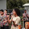 Uma mulher canta ao microfone enquanto uma banda toca instrumentos de percussão ao fundo, durante uma celebração cultural ao ar livre.