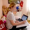 Uma jovem de cabelos loiros presos em um rabo de cavalo está sentada em uma cama em um quarto, usando um computador portátil enquanto escreve em um caderno.