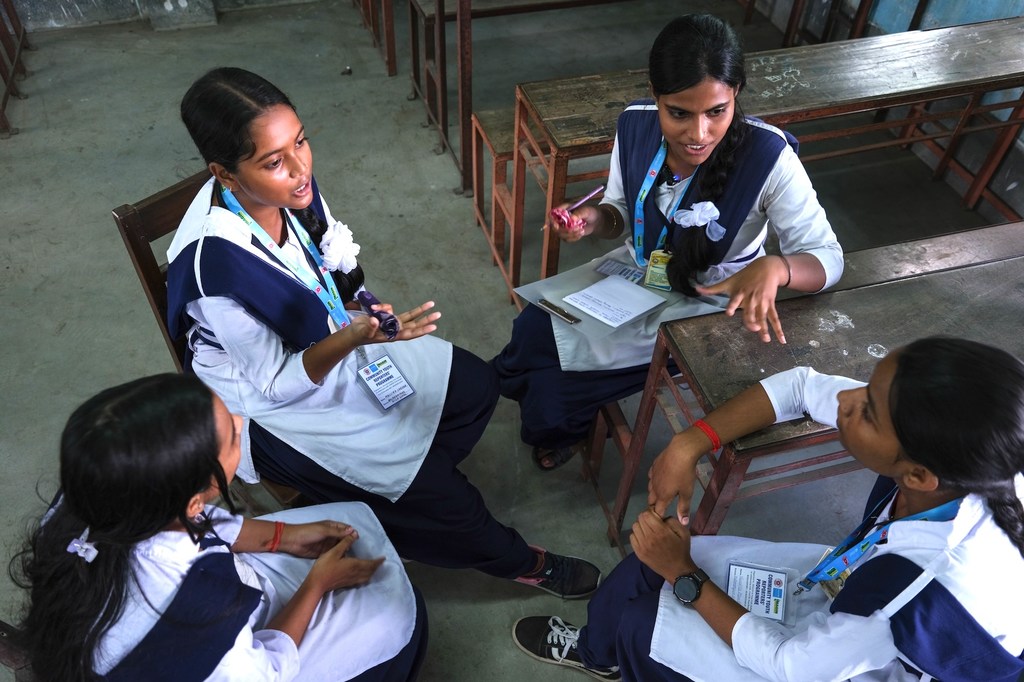 स्कूल यूनिफॉर्म पहने चार छात्राएं कक्षा में बैठी हैं और एक कहानी के विचार पर चर्चा कर रही हैं। एक छात्रा के हाथ में पेन और एक छोटी नोटबुक है, जबकि अन्य छात्राएं सुन रही हैं और बातचीत में भाग ले रही हैं।