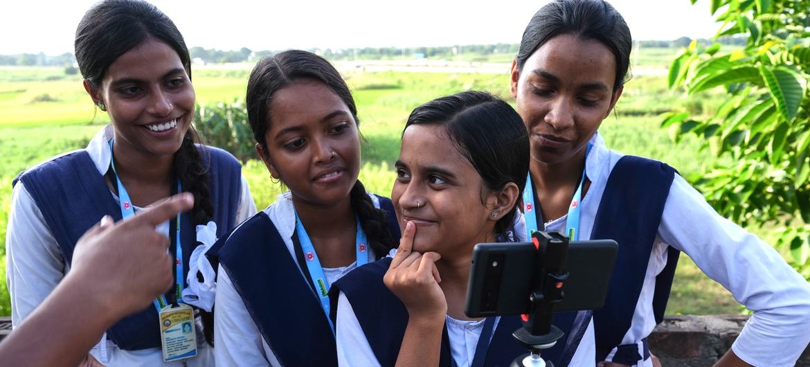 यूनिफॉर्म पहने चार युवतियां बाहर इकट्ठा होकर गिम्बल पर लगे स्मार्टफोन का इस्तेमाल करना सीख रही हैं। वे पश्चिम बंगाल के मुर्शिदाबाद में UNICEF इंडिया के युवा रिपोर्टर कार्यक्रम का हिस्सा हैं।