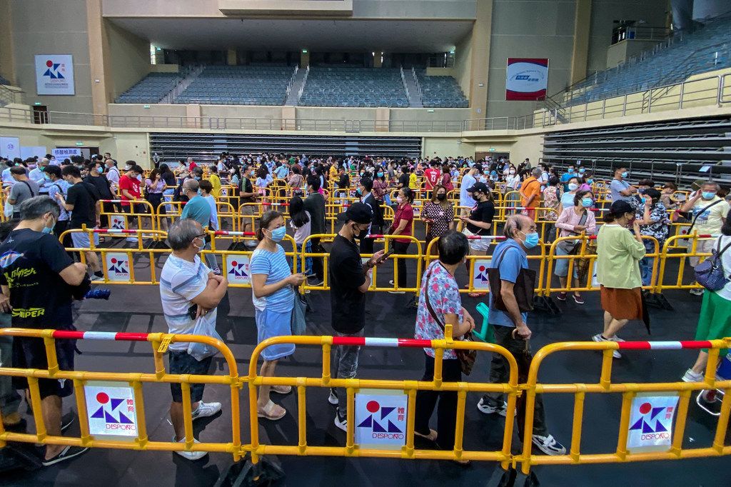 Multitudes de personas con mascarillas hacen cola dentro de un gran pabellón deportivo cubierto para someterse a pruebas masivas de COVID-19 en Macao.