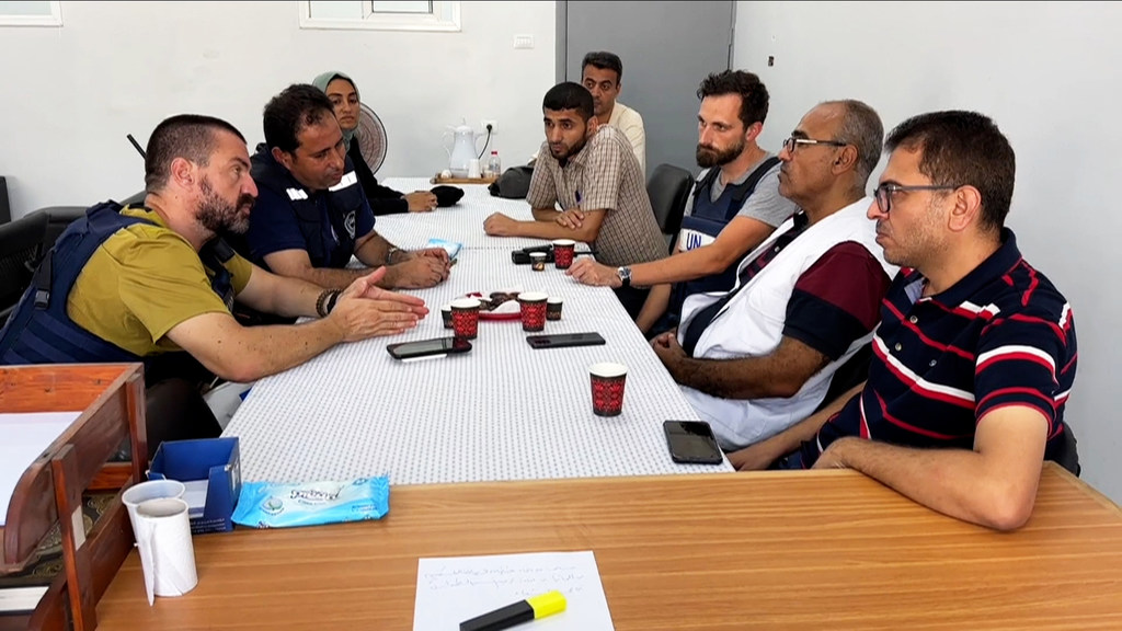 مجموعة من الرجال وامرأة واحدة يرتدون سترات الأمم المتحدة يجلسون حول طاولة مؤتمرات في غرفة اجتماعات، منخرطين في نقاش.