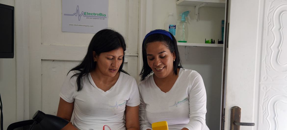 Duas mulheres venezuelanas trabalham nos componentes internos de um computador em uma oficina, uma delas segurando um multímetro amarelo.