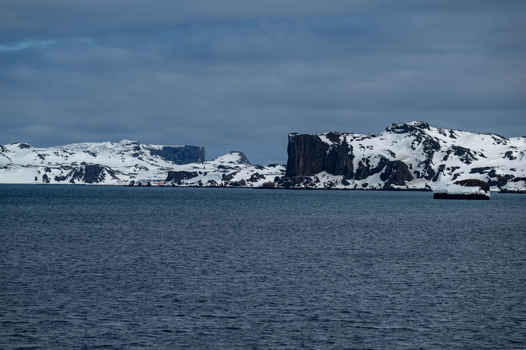 Vista de montañas rocosas cubiertas de nieve en la Península Antártica, vistas al otro lado de una masa de agua oscura bajo un cielo nublado.
