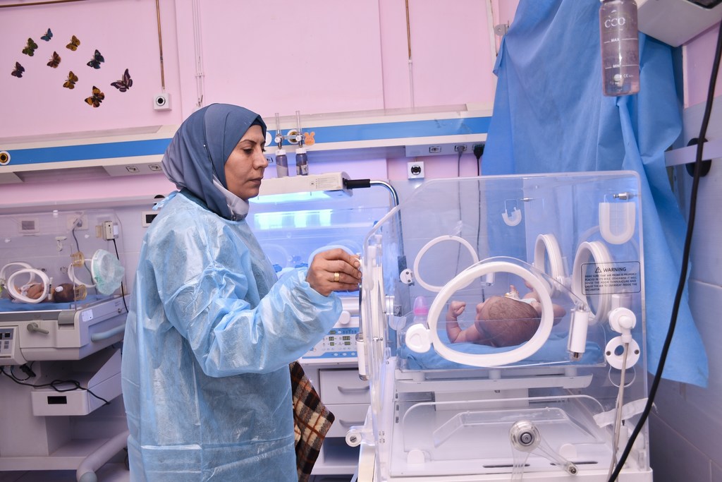 تقف ممرضة ترتدي ملابس واقية بجوار حاضنة تحتوي على طفل حديث الولادة في وحدة العناية المركزة لحديثي الولادة.