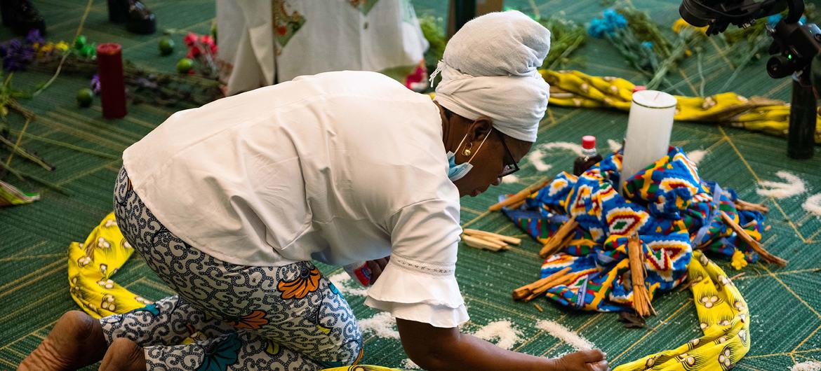 Una mujer africana con un turbante blanco se arrodilla sobre una alfombra verde, acomodando telas de colores para una ceremonia en la Cumbre Nacional de Mujeres y Paz en Bogotá.