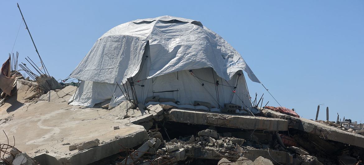 Un gran refugio de lona blanca se alza sobre un montón de escombros y restos bajo un cielo azul despejado.