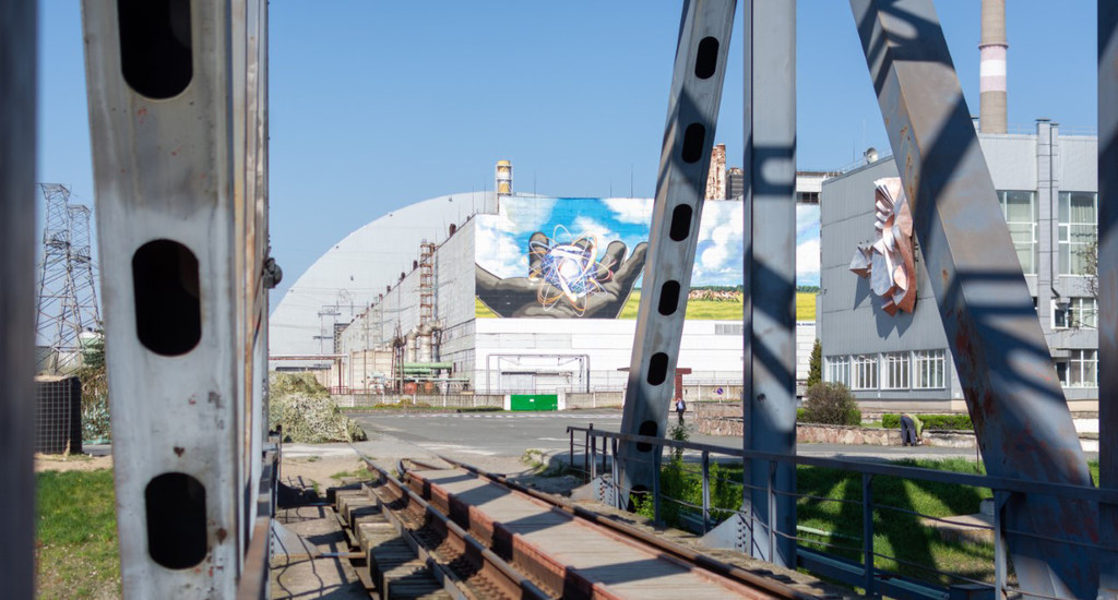 Вид на Чернобыльскую атомную электростанцию в Украине, где изображена массивная конструкция нового безопасного укрытия с большой фреской рук, держащих атом. На переднем плане под чистым голубым небом видны железнодорожные пути и промышленные металлические конструкции.