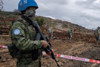 छलावरण वाली वर्दी और नीले रंग का UN का हेलमेट पहने चीनी UNIFIL लड़ाकू इंजीनियर अग्रभूमि में खड़ा है, जबकि अन्य सैनिक पृष्ठभूमि में एक बारूदी सुरंग हटाने वाले स्थल पर काम कर रहे हैं।