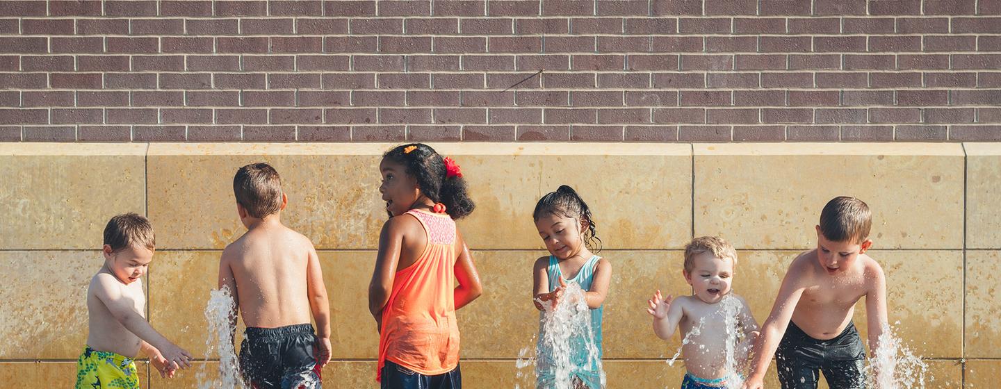 Plusieurs enfants jouent dans une fontaine, avec des murs de briques et de pierres en arrière-plan.