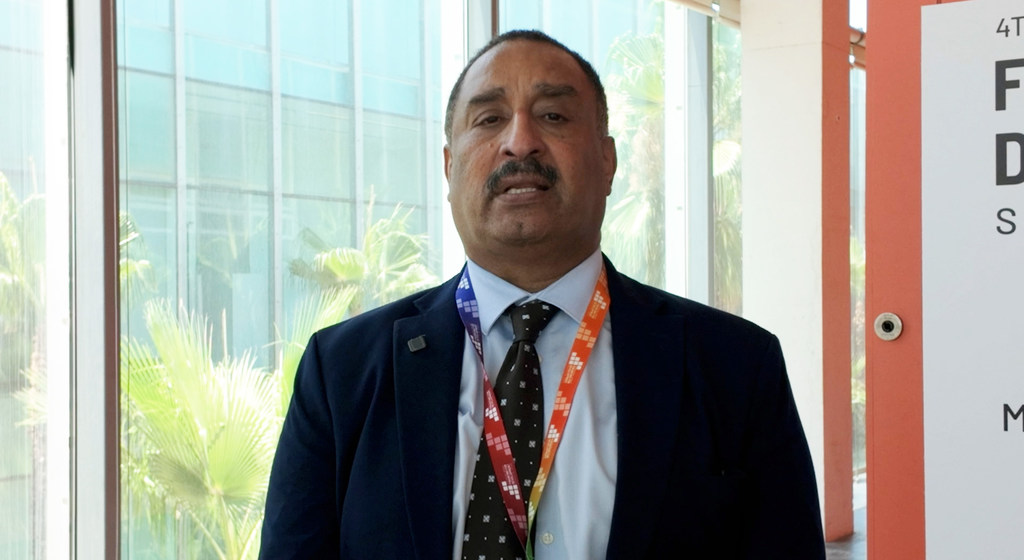 يظهر هاشم حسين، رئيس مكتب منظمة الأمم المتحدة للتنمية الصناعية - منظمة التجارة الدولية في البحرين، في الصورة مرتدياً بدلة مع حبل تعليق، أمام جدار زجاجي.