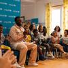 Um homem negro está sentado em uma sala de aula em Portugal, rodeado por um grupo diversificado de crianças e adultos que aplaudem durante um evento da Unicef .