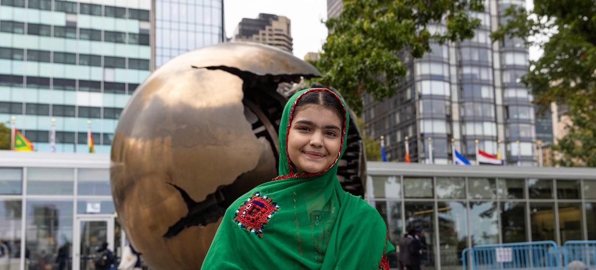 سبز کڑھائی والی شال میں ایک نوجوان عورت شہری پلازہ میں ایک بڑے، پھٹے ہوئے گلوب مجسمے کے سامنے کھڑی ہے۔