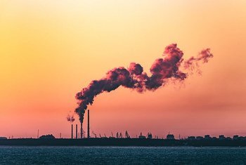 صورة ظلية لمداخن صناعية تنبعث منها دخان داكن على خلفية سماء غروب الشمس الملونة فوق مسطح مائي.
