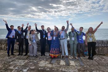 A group of diverse young participants at the UN Alliance of Civilizations Youth Forum in Cascais, Portugal, waving and smiling by the seaside on November 25, 2024.