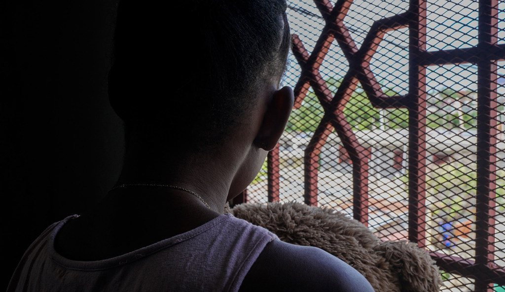 En Haití, se ve a un niño pequeño de espaldas, sosteniendo un osito de peluche mientras mira por una ventana con barrotes de seguridad.