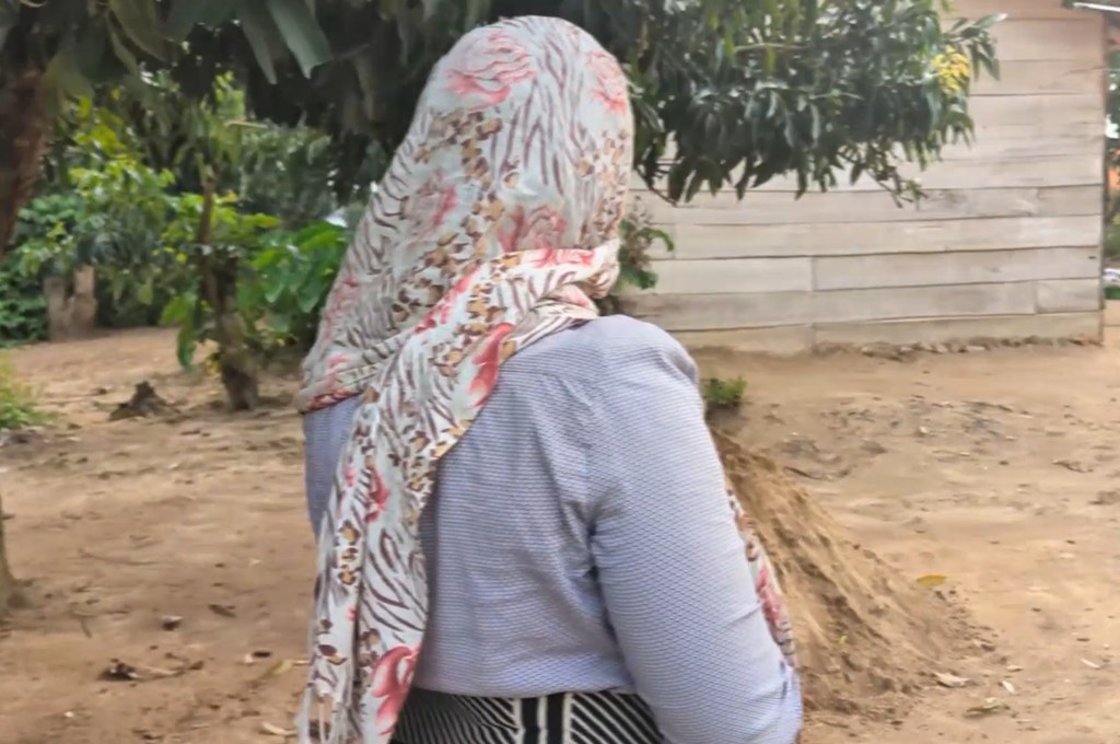 Une femme portant un foulard à fleurs et un haut gris se tient dos à la caméra dans un décor rural.
