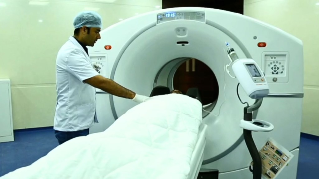 Un professionnel de santé, vêtu d'une blouse blanche et d'une charlotte chirurgicale, manipule un appareil d'IRM et guide un patient recouvert d'un drap blanc à l'intérieur de la machine.