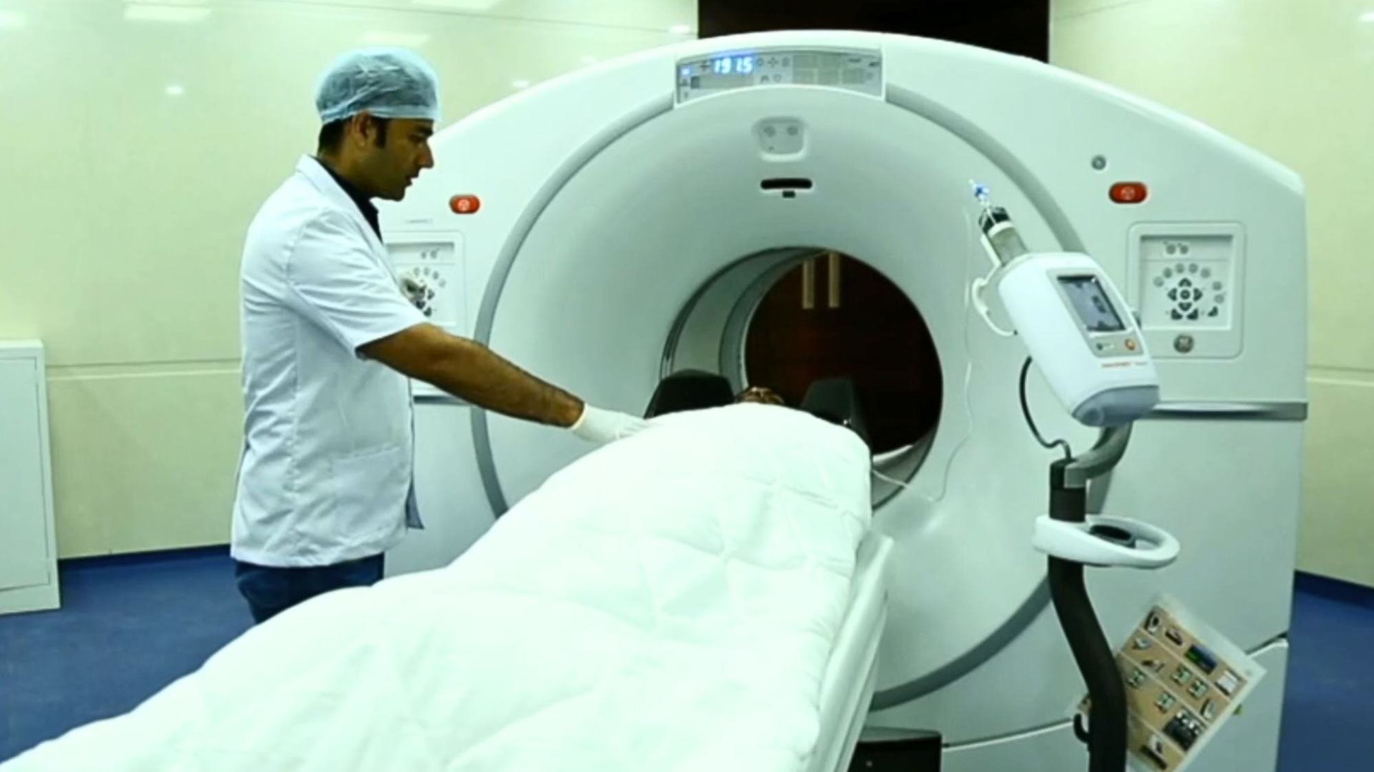 A male medical professional in a white coat and surgical cap operates an MRI scanner, guiding a patient covered with a white sheet into the machine.