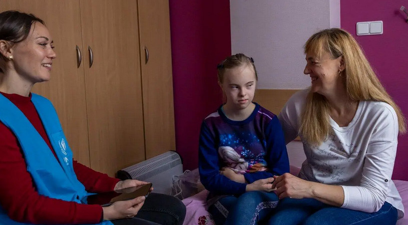 Волонтер UNHCR в синем жилете сидит дома с матерью и ее дочерью, страдающей синдромом Дауна.