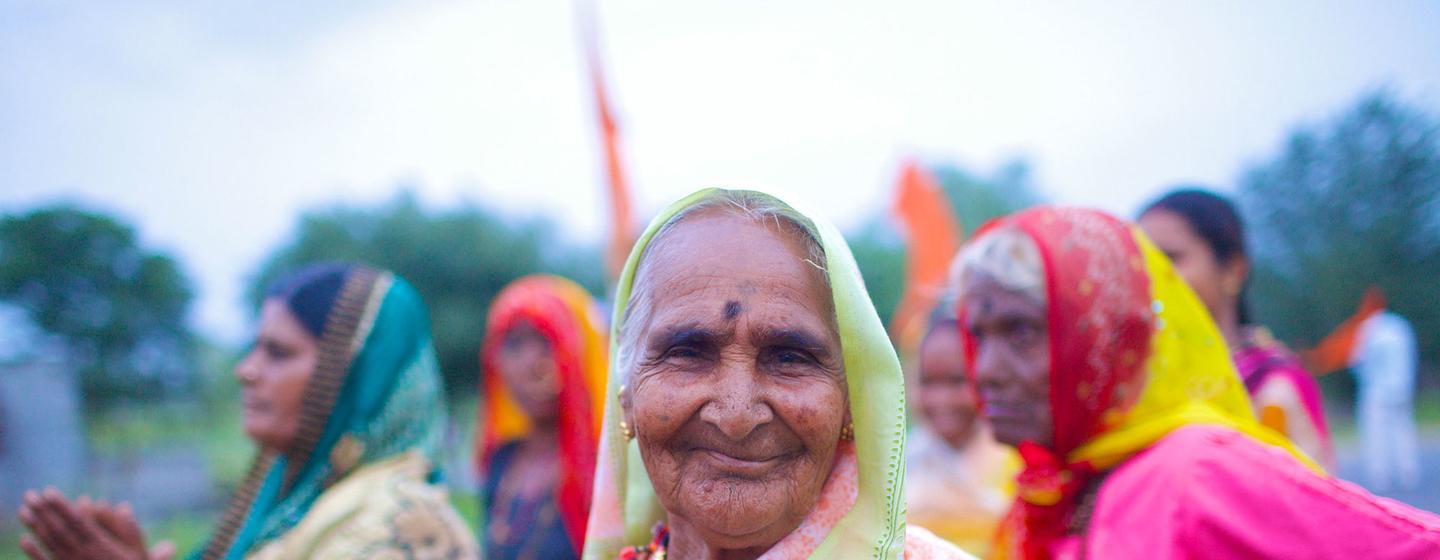 Une femme indienne âgée, vêtue d'un sari, sourit, les mains jointes en signe de prière, avec d'autres femmes en arrière-plan.