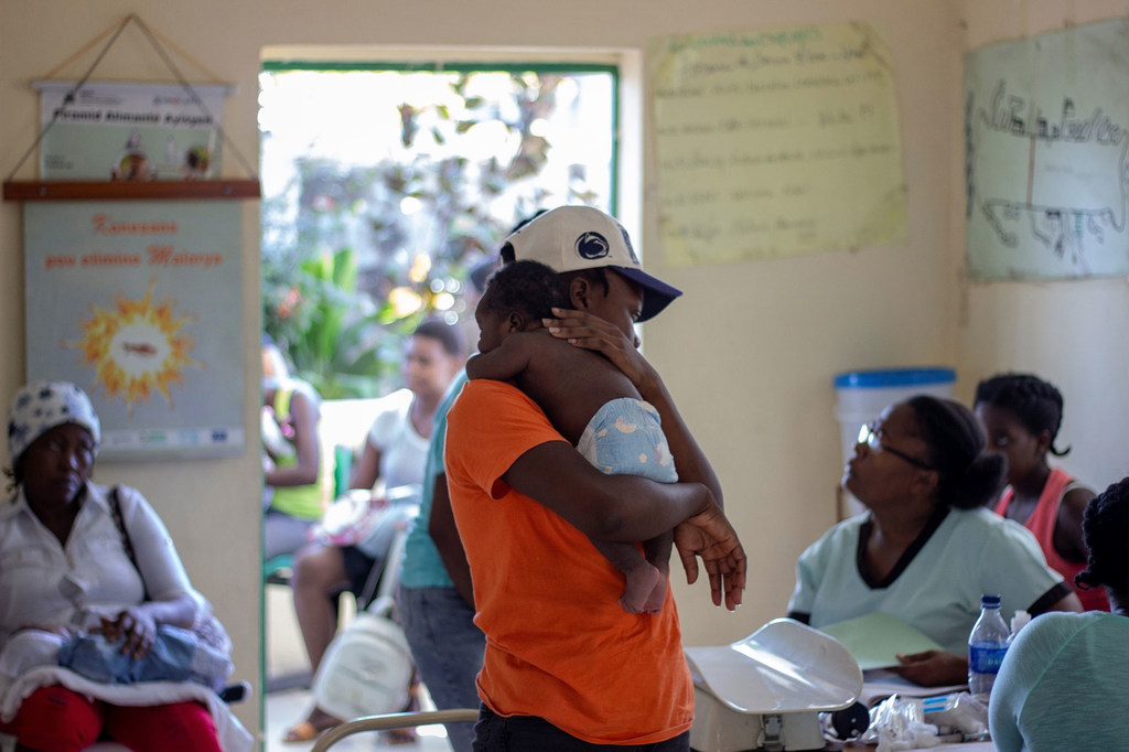 Um homem com camisa laranja e boné de beisebol segura um bebê em uma clínica no Haiti, enquanto outras pessoas aguardam nas proximidades.