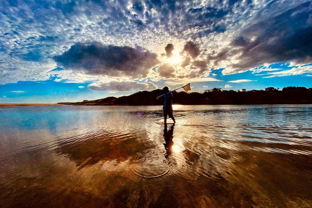 Um pescador está em águas rasas segurando uma rede, com um céu de pôr do sol dramático e nuvens refletidas na superfície.