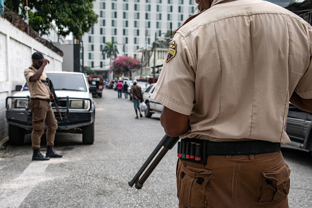 A Haitian National Police officer stands on a street in Port-au-Prince, with other police and civilians in the background.