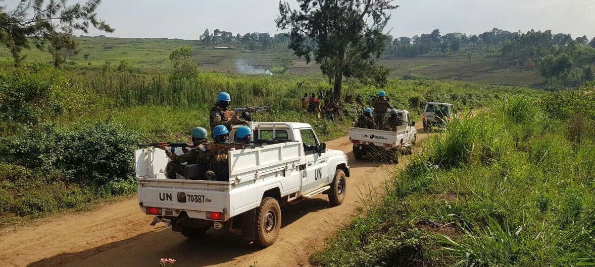 Soldados de paz da ONU em caminhonetes brancas patrulham uma estrada de terra em uma paisagem rural.