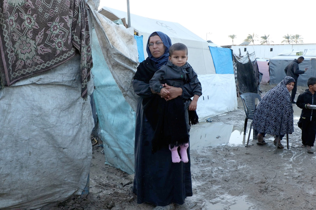 امرأة فلسطينية تحمل طفلاً صغيراً أمام خيمة متضررة من المياه في مخيم الكرامة للنازحين في غزة.