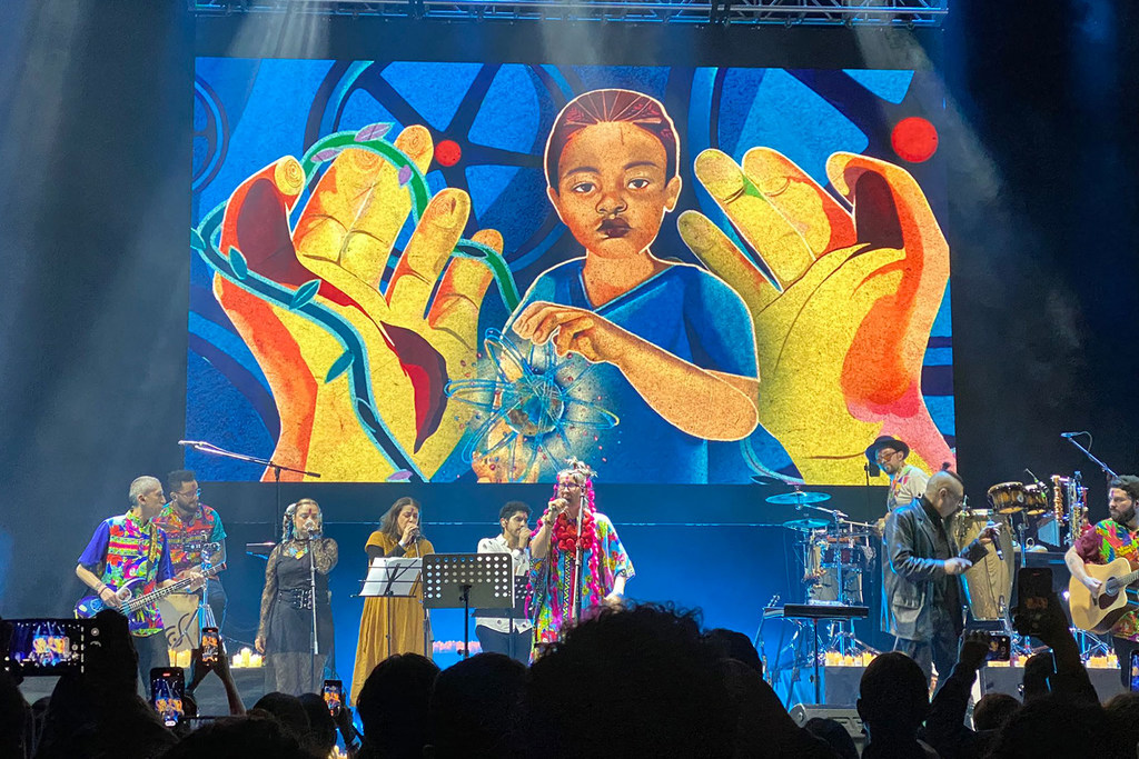 Los músicos Aterciopelados, Susana Baca, Enrique Bunbury y el Dr. Shenka actúan en un escenario frente a una gran pantalla que muestra la imagen de las manos de un niño.