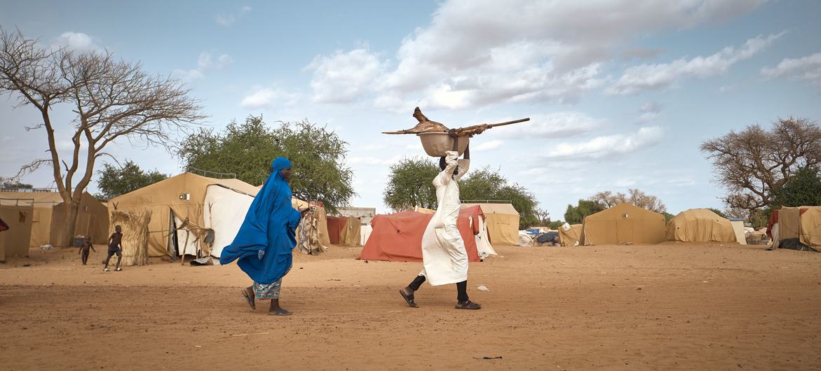 Duas pessoas caminham por um campo de deslocados em Niamey, Níger, sob um céu azul. Uma pessoa vestindo uma túnica azul caminha ao lado de outra vestida com uma túnica branca, que carrega uma grande panela na cabeça.