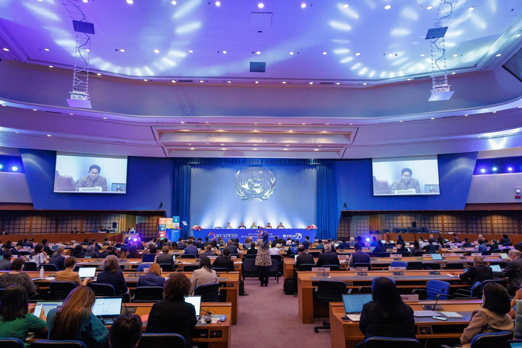 United Nations की एक बैठक में एक विशाल सम्मेलन कक्ष, जिसमें एक मंच, बड़ी स्क्रीन और कई प्रतिभागी डेस्क पर बैठे हुए हैं।