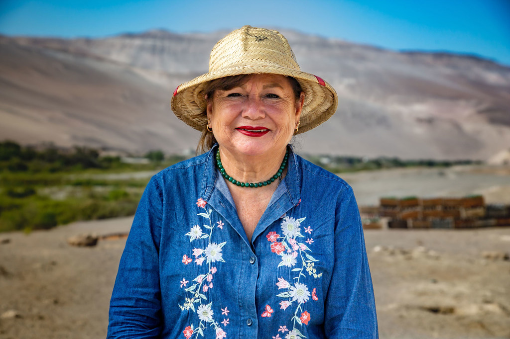 Retrato de Edith Elgueta, apicultora chilena, con sombrero de paja y camisa azul bordada, con un paisaje desértico de fondo.